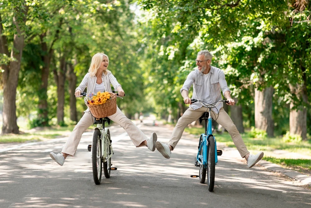 Senior couple joyfully riding bicycles together on a sunny path, with a basket of flowers on the woman’s bike.